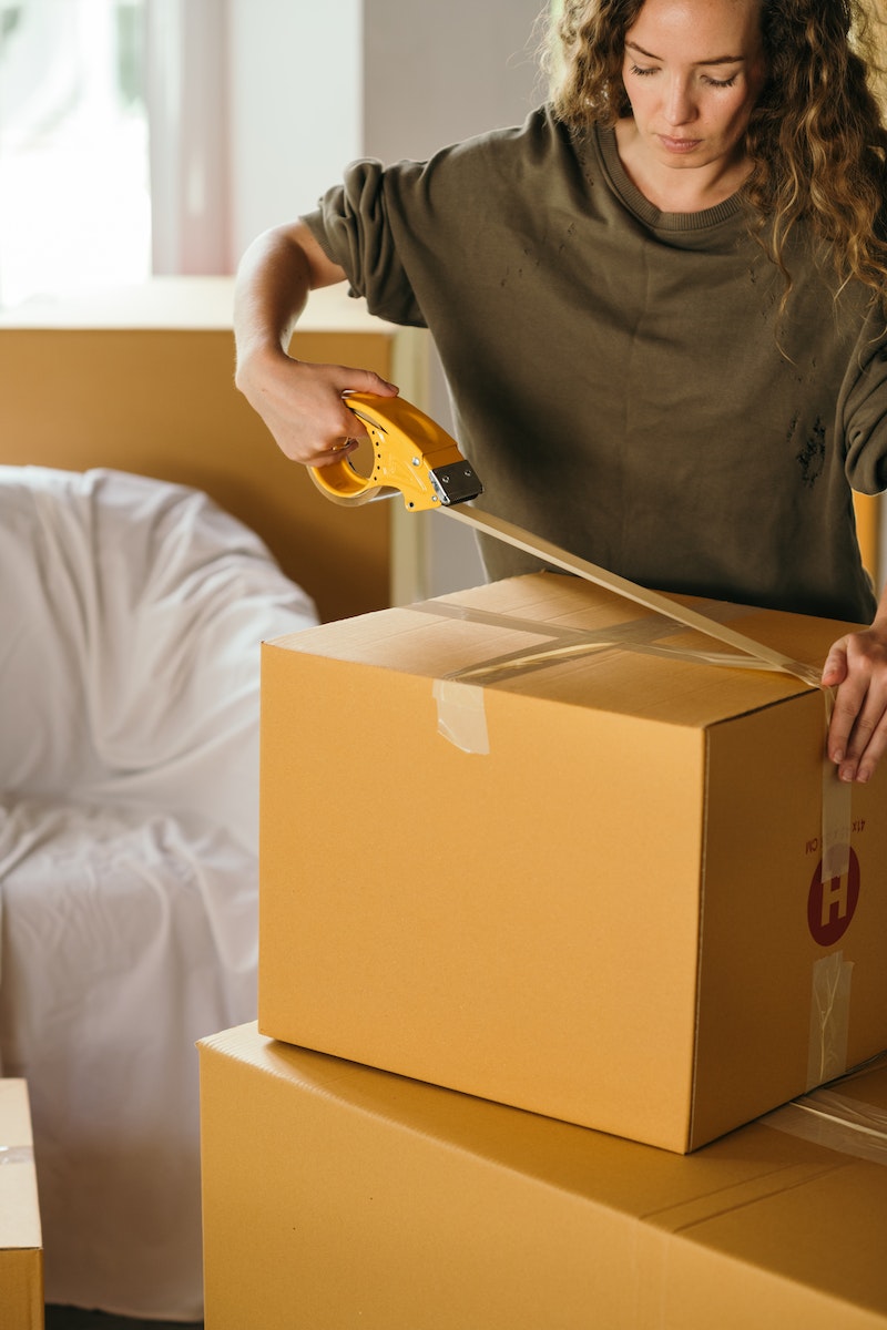 moving abroad checklist, Crop female wearing casual clothes standing while packing carton boxes with scotch tape in modern apartment during preparing for relocation, moving abroad checklist